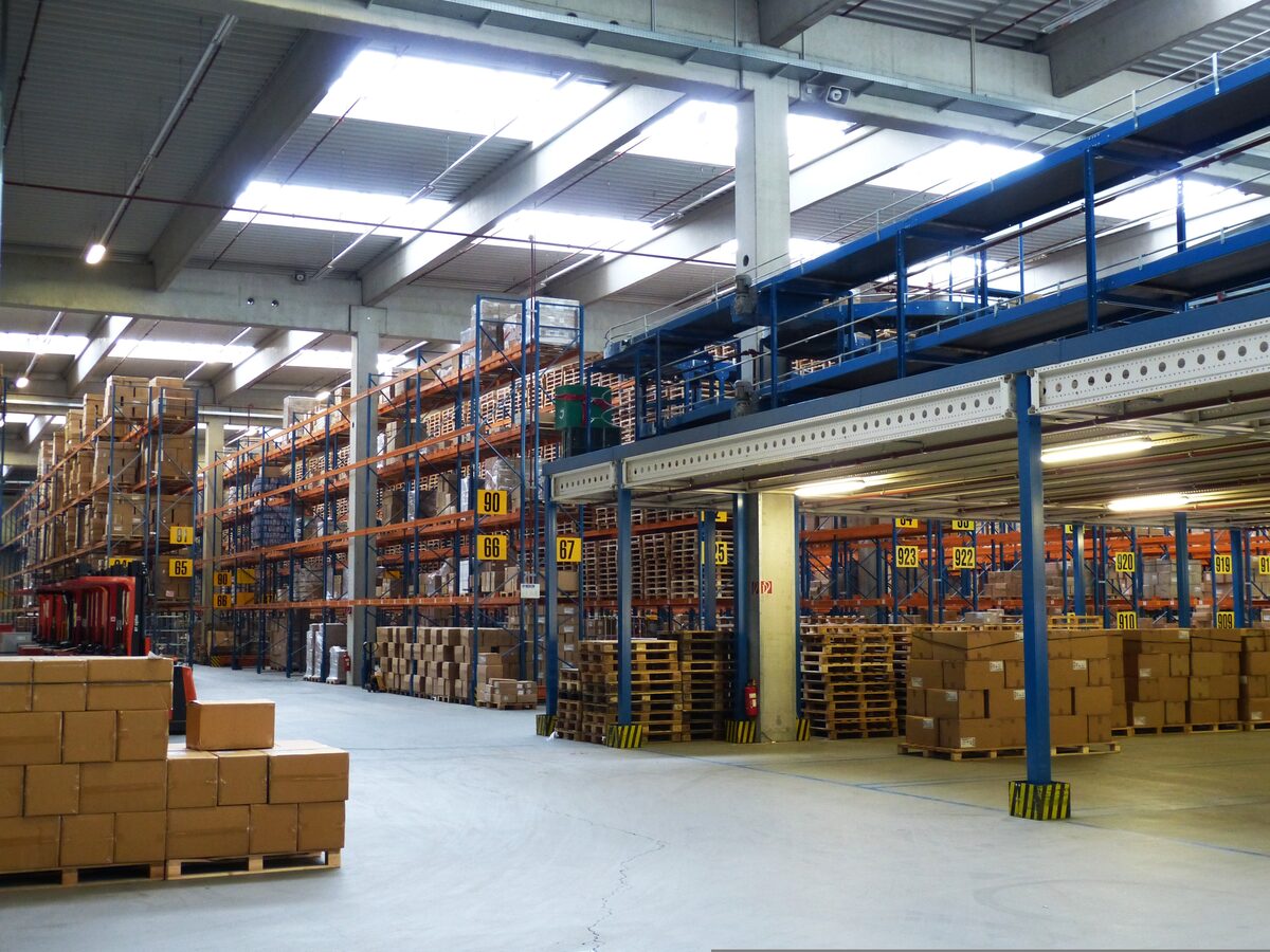 Interior of a high-bay distribution warehouse — towering pallet racks of brand-authorized inventory organized by numbered bay for nationwide retail fulfillment.